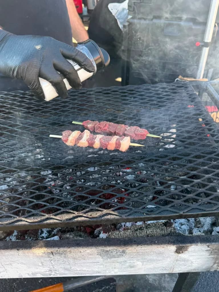 A cook seasoning beef skewers over glowing coals